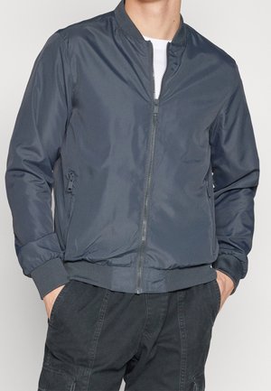 Homme portant une veste bomber zippée gris foncé avec poignets, col et ourlet côtelés, debout les mains dans les poches d’un pantalon cargo noir.