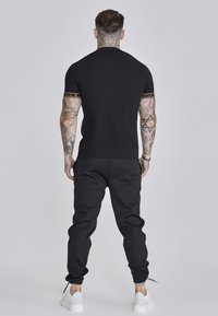 Sort tætsiddende t-shirt med guld logo akcenter på ærmerne, parret med matchende sorte sweatpants og hvide sneakers. Simpelt, atletisk design.