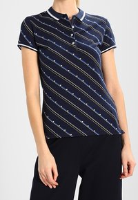 Polo blu navy con bordi bianchi, caratterizzato da strisce diagonali e piccoli motivi a cuore. Maniche corte e bottoni al colletto.