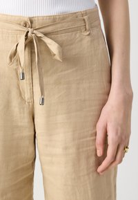 Beige linen pants with a drawstring waist, featuring metallic tips on the ties and side pockets. Smooth texture and loose fit.