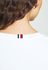 Zbliżenie na szyję i ramię osoby noszącej białą koszulę z małą czarno-biało-czerwoną metką z tyłu kołnierzyka.