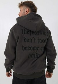 Donkergrijze hoodie van een katoenmix, met grote zwarte tekst op de achterkant. Het heeft een buidelzak en ribgebreide cuffs en zoom.