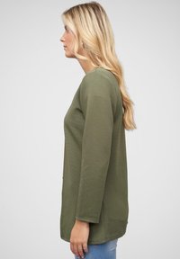 Olive green long-sleeve top with a textured ribbed pattern, featuring a relaxed fit and side pockets, paired with light denim jeans.