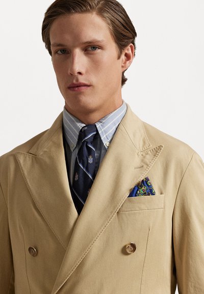 Beżowa podwójnie zapinana marynarka z dużymi klapami, noszona na niebieskiej i białej w paski koszuli oraz ciemnym krawacie z wzorem. Poszetka dodaje koloru.