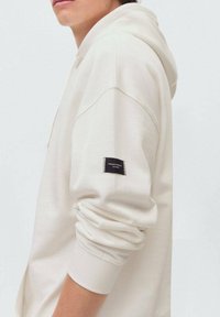 Krämfärgad hoodie med en avslappnad passform, som har en dragsko i huvan och ribbade ärmslut. Svart etikett på vänster ärm. Mjuk textur.