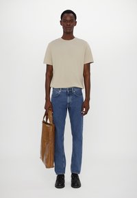 Camiseta de algodón color beige, jeans de mezclilla azules y zapatos negros. Bolsa de mano marrón con un acabado brillante y sección transparente, sostenida con la mano izquierda.
