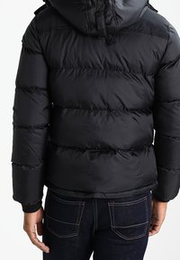 Black puffer jacket with a hood, featuring horizontal quilted sections, adjustable drawstring, and elastic cuffs, worn over dark jeans.