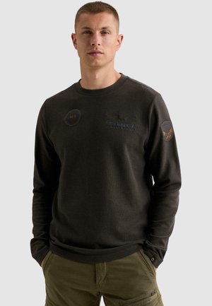 Homme portant un sweat-shirt à manches longues foncé avec des patches sur la poitrine et la manche, et un pantalon cargo vert olive avec les mains dans les poches.