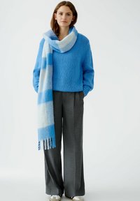 Pull en maille texturée bleu avec des manches larges, associé à un pantalon tailleur gris. Une écharpe à carreaux bleu clair et blanc se drape autour du cou.