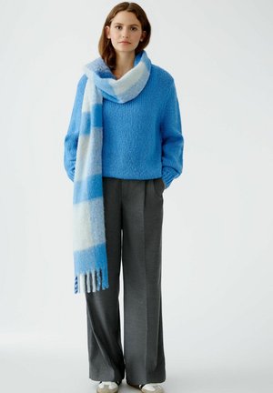 Pull en maille texturée bleu avec des manches larges, associé à un pantalon tailleur gris. Une écharpe à carreaux bleu clair et blanc se drape autour du cou.