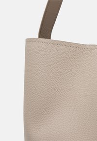 Gros plan d’un sac à main en cuir beige texturé avec des coutures visibles et une seule sangle sur un fond clair uni.