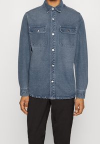 Person, der ein blaues Denim-Hemd mit Druckknöpfen und zwei Brusttaschen trägt, kombiniert mit schwarzen Hosen.