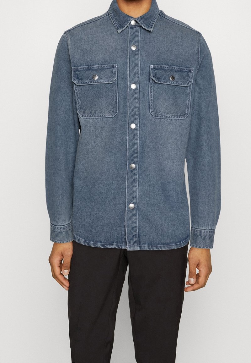 Person, der ein blaues Denim-Hemd mit Druckknöpfen und zwei Brusttaschen trägt, kombiniert mit schwarzen Hosen.