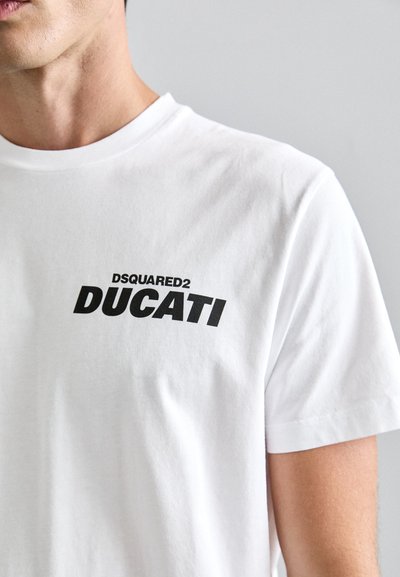 Valge puuvillane T-särk, millel on ümar kaelus, lühikesed varrukad ja julge must tekst: "DSQUARED2 DUCATI" ees. Sile kangastruktuur.