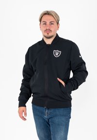 Giacca bomber nera con zip realizzata in tessuto morbido, con logo dei Raiders sul petto e polsini e orlo a coste. Include tasche laterali.