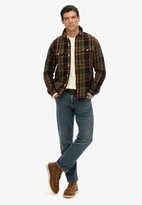 Geruite blouse in donkere tinten met oranje accenten, gedragen over een witte trui, gecombineerd met blauwe jeans en bruine veterschoenen.