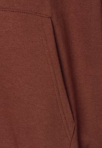 Brun hettegenserstoff med en teksturert overflate og en sidelomme. Synlig stitching langs kantene. Myk og uformell design.