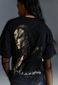 Černé tričko s grafickou potiskem bledého obličeje s tmavými vlasy, oblékajícího texturovaný plášť, a textem "Frankenstein" v bílé barvě.