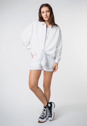 Weißes gemustertes Hemd mit Knöpfen, in der Taille gebunden, kombiniert mit weißen Shorts mit gewelltem Saum, ergänzt durch schwarze High-Top-Sneaker.