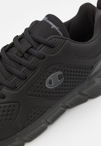 Schwarzer Sportschuh mit strukturierter Oberfläche, ausgestattet mit Schnürsenkeln, einer gepolsterten Zunge und einer Gummisohle. Logo in Grau an der Seite.