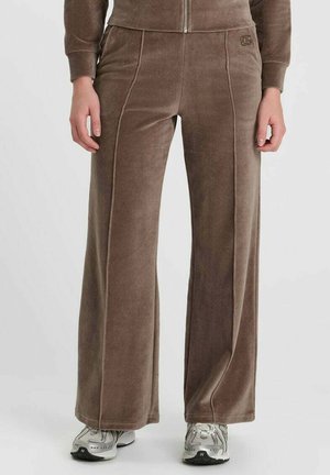 Pantaloni larghe in velluto marrone indossati con sneakers argento e bianche, visibili dalla vita ai piedi su uno sfondo semplice.
