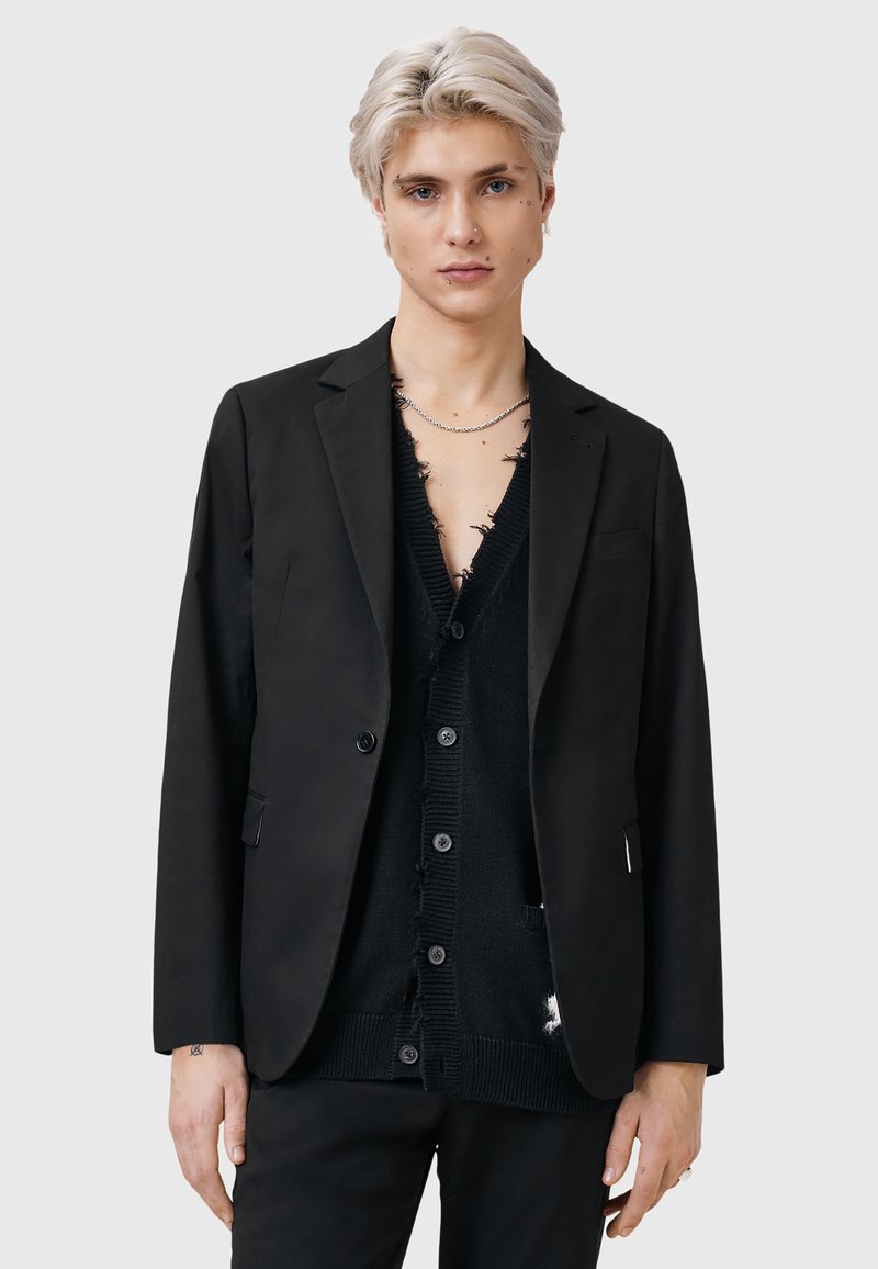 Svart blazer i slät tyg med slagkrage och två knappar, bärs över en sliten svart cardigan med djup V-hals och knäppning.