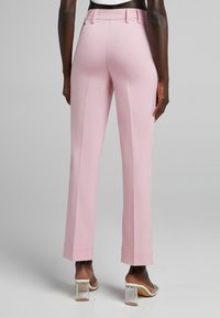 Osoba v světle růžových rovně střižených kalhotách a sandálech s průhledným podpatkem, stojící na pozadí v jemně šedé barvě.