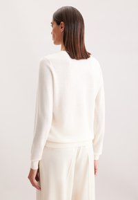 Maglione bianco lavorato a maglia con maniche lunghe, polsini e orlo a coste, e una consistenza morbida, indossato con pantaloni color crema. Vista da dietro.