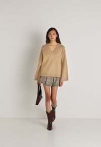 Mulher vestindo um suéter bege de V, saía mini plissada xadrez, botas castanhas e segurando uma pequena mala escura, caminhando sobre um fundo liso.