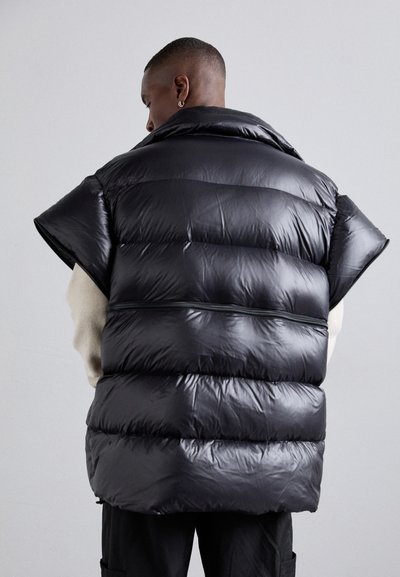 Gilet en duvet noir avec une finition brillante, emmanchures larges et sections matelassées horizontales. Il dispose d'un large col et d'un zip dans le dos.