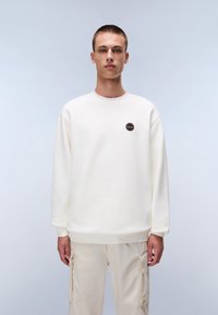 Sudadera blanca de cuello redondo con mangas largas, que presenta un pequeño parche redondo. Tejido suave con puños y dobladillo acanalados, combinado con pantalones cargo beige.
