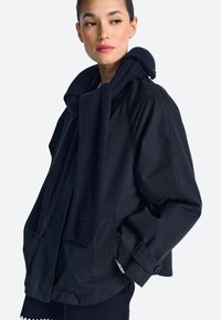 Tamsiai mėlyna striukė su struktūrizuotu šaliku, laisvo silueto, su didelėmis kišenėmis ir užsegamais rankogaliais. Audinys atrodo lygus ir patvarus.