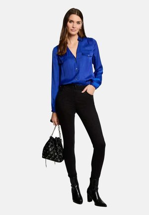 Camicia blu in satin con bottoni e due tasche sul petto, abbinata a jeans skinny neri e stivali alla caviglia. Porta una borsa a tracolla nera con motivo.