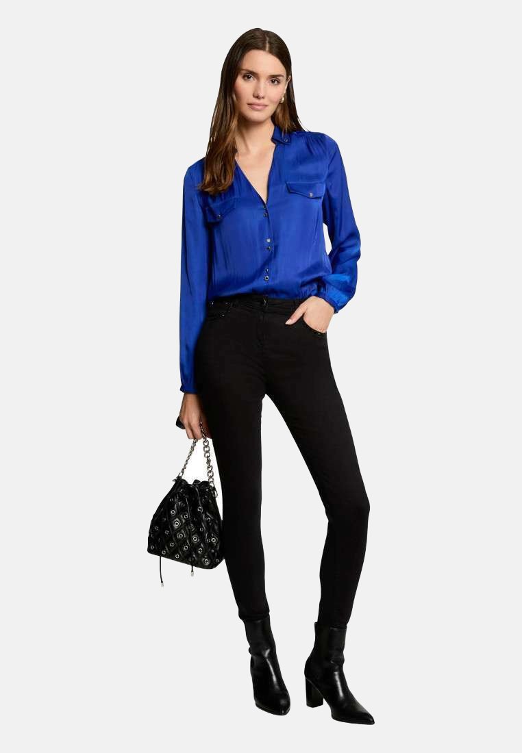 Camicia blu in satin con bottoni e due tasche sul petto, abbinata a jeans skinny neri e stivali alla caviglia. Porta una borsa a tracolla nera con motivo.