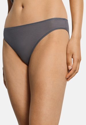 Frau trägt schlichte graue Bikini-Unterwäsche mit niedrigem Bund, die den Bauchbereich und die oberen Oberschenkel vor neutralem Hintergrund zeigt.