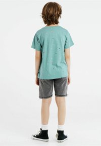 Lapsi, jolla on vaaleanruskeat hiukset, pukeutuneena teal-väriseen printtipaitaan, harmaisiin shortsseihin, valkoisiin sukkiin ja mustiin korkeisiin tennareihin, seisoo selkä kameraan päin.