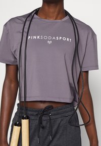 Person som bär en kort lila Pink Soda Sport-tröja och mörkgrå byxor med dragsko, håller i ett hopprep med trähandtag.