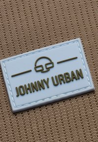 Světle modrá gumová etiketa s vyraženým tmavě zeleným textem "JOHNNY URBAN" a ikonou brýlí, ohraničená šitou bílou hranou na hnědém textilu.