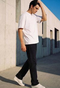 Young man in white t-shirt and black pants walking beside a concrete building, touching his hair with his right hand.