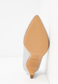 Chaussure à talon haut pailleté argent à bout pointu, reposant semelle vers le haut contre un mur blanc, montrant une semelle beige et la taille 38.