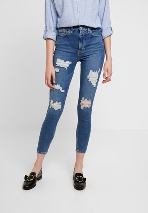 Frau trägt hoch taillierte, enge blaue Jeans mit mehreren großen Rissen, kombiniert mit schwarzen Loafers mit goldenen Schnallen und einem hellblauen Hemd mit hochgekrempelten Ärmeln.