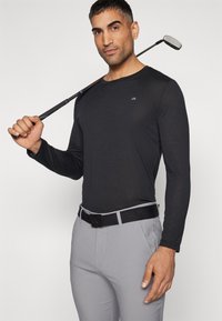 Svart langermet skjorte med CK-logo, kombinert med lysegrå bukser og et svart belte, holder en golfkølle over skulderen. Glatt tekstur.
