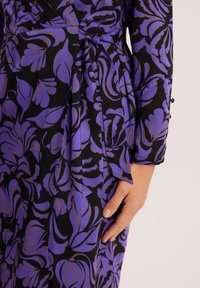 Fioletowa sukienka w kwiatowy wzór na czarnym tle, z zapinanym krojem, wiązanym w pasie i długimi rękawami. Tkanina ma gładką teksturę.