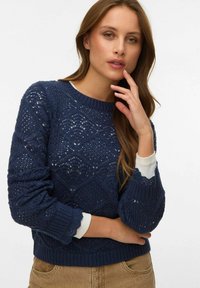 Femme aux longs cheveux bruns portant un pull en maille texturée bleu marine et un pantalon beige, posant avec la main près du visage.