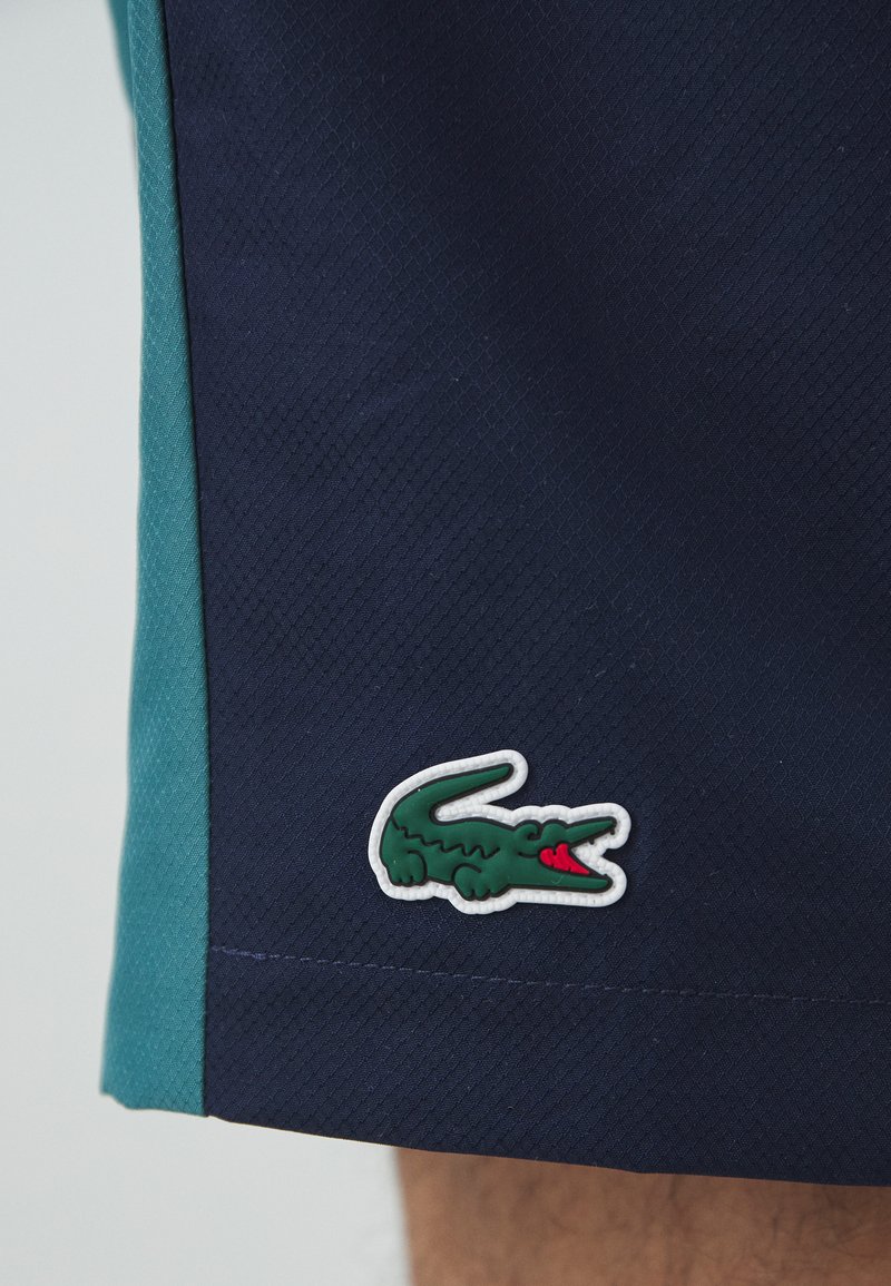 Shorts navy avec des panneaux latéraux turquoise, en tissu texturé, avec un logo en contour blanc représentant un crocodile vert à la bouche rouge.