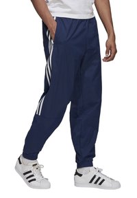 Muž v tmavě modrých teplákových kalhotech s bílými bočními pruhy a bílých teniskách Adidas s černými pruhy, stojící s jednou rukou v kapse.