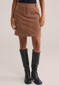 Jupe mini en velours côtelé marron avec une silhouette droite et une taille ; accompagnée de hautes bottes en cuir noir avec une fermeture éclair sur le côté.