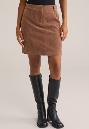 Jupe mini en velours côtelé marron avec une silhouette droite et une taille ; accompagnée de hautes bottes en cuir noir avec une fermeture éclair sur le côté.