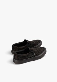 Un par de zapatillas negras sin cordones con un sutil patrón de leopardo y suelas de goma sobre un fondo blanco.