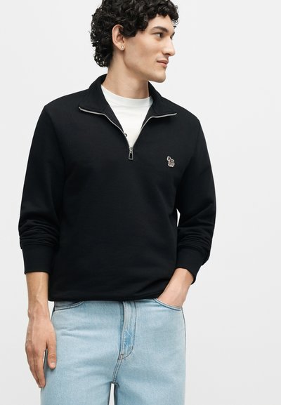 Νέος άνδρας με μαύρο φούτερ με φερμουάρ μέχρι τον λαιμό και ένα μικρό κεντητό πατς με ζέβρα, ανοιχτό μπλε τζιν και λευκό πουκάμισο από μέσα, όρθιος.